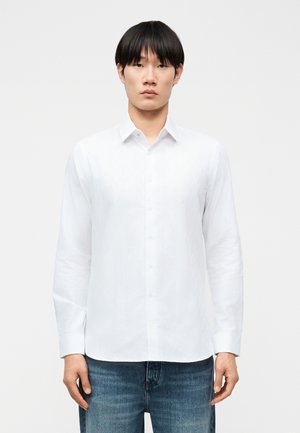 Młody mężczyzna o czarnych włosach, ubrany w białą koszulę na guziki i niebieskie dżinsy, stojący na tle jednolitego białego tła.