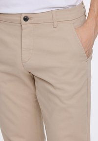 Bēšas krāsas bikses ar glītu taisnu piegriezumu, plakanu priekšpusi, sānu kabatām un vienu pogu aizdares vietā. Auduma tekstūra ir gluda un izturīga.