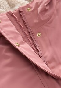 Primer plano de una chaqueta rosa con cremallera de plástico parcialmente abierta y botones de presión dorados, mostrando un forro beige suave en el interior del cuello.