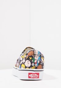 Chaussure slip-on en toile avec un motif coloré de personnages Peanuts, fond noir, semelle en caoutchouc blanche et logo Vans au talon.