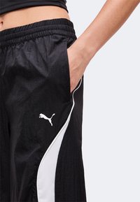 Κοντινό πλάνο ατόμου που φορά μαύρο φόρμα Puma με λευκές πλευρικές ρίγες, χέρι στην τσέπη και ορατή ελαστική μέση.