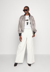 Metāliskā sudraba bumbierjakete, balts grafiskais T-krekls ar varoņa ilustrāciju, platai baltās bikses un melni smailu purngalu zābaki.