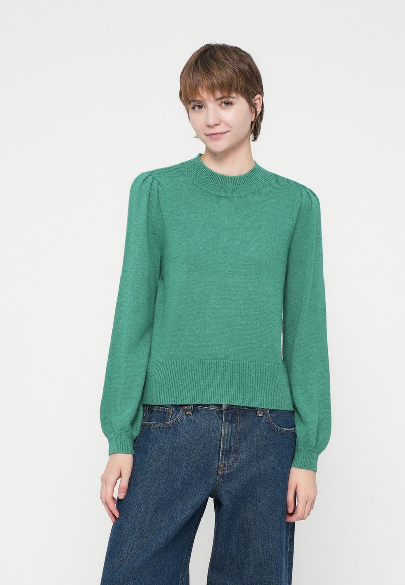 Giovane che indossa un maglione verde a maniche lunghe e jeans scuri larghi, in piedi contro uno sfondo bianco uniforme.