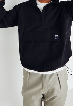 Person trägt eine schwarze Levi's-Pulloverjacke über einem weißen Hemd mit beigen Hosen, Arme aus dem Bildrahmen erhoben vor einem schlichten Hintergrund.