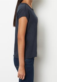 Donna in piedi di lato, che indossa una maglietta scura a maniche corte e jeans blu, davanti a uno sfondo bianco semplice.