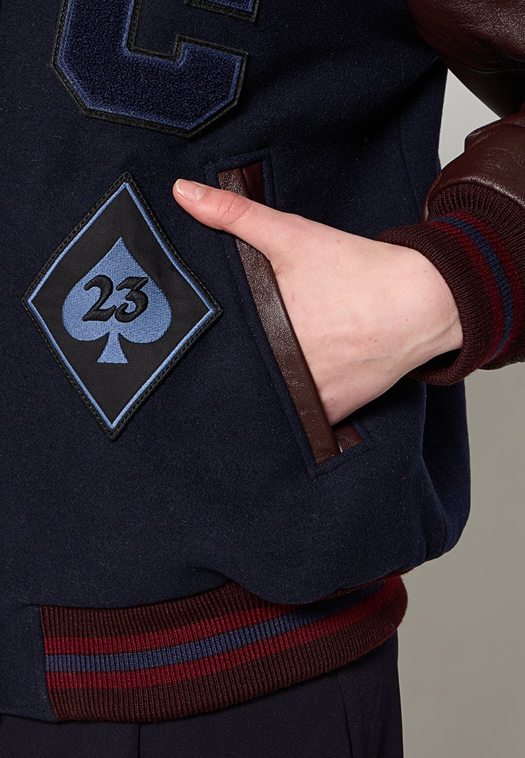 Giacca di lana navy con dettagli in pelle marrone, caratterizzata da una toppa a forma di diamante con un asso blu e il numero 23, polsini a coste in color bordeaux e blu.