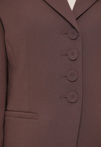 Hnedé saka na mieru z textúrovanej látky s vykrojeným klopou, štyrmi kruhovými gombíkmi a bočným vreckom.