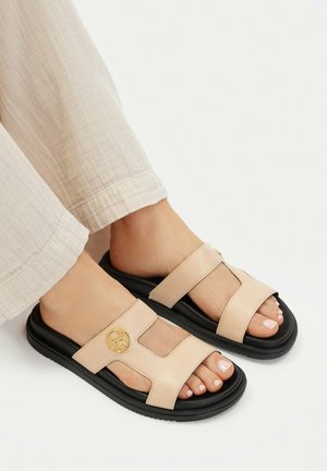 Piedi che indossano sandali beige con punta aperta e suole nere con dettaglio logo dorato, abbinati a pantaloni beige chiaro su sfondo bianco.