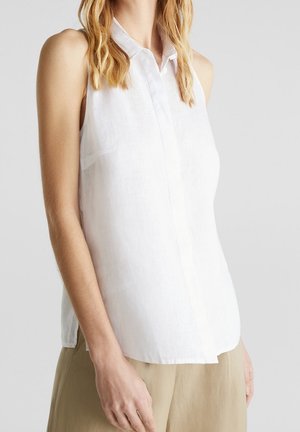 Blouse blanche sans manches à col avec boutons, portée avec un pantalon beige, présentée sur une personne debout sur un fond clair uni.