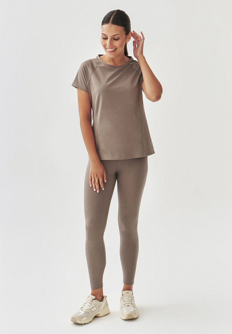 Top corto a maniche corte marrone e leggings coordinati realizzati in un tessuto liscio e elasticizzato. La modella indossa scarpe da ginnastica chiare. Design semplice.