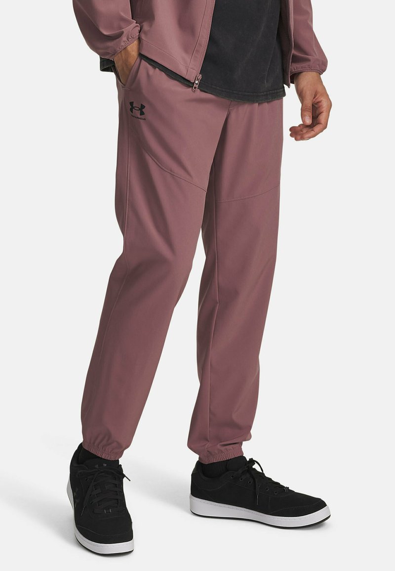 Licht mauve joggerbroeken van gladde stof, met elastische boorden, zijzakken en een subtiele logo op de linker bovenbeen. Zwarte sneakers zijn afgebeeld.