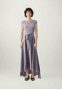 Woman standing wearing a sleeveless lavender lace top dress with a tied waist and asymmetrical hem, paired with black open-toe heels.