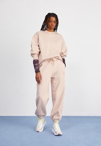 Ung person som står, klädd i en lös beige sweatshirt och sweatpants, lila mönstrade långärmade tröja under sweatshirten, vita sneakers med neonaccenter.