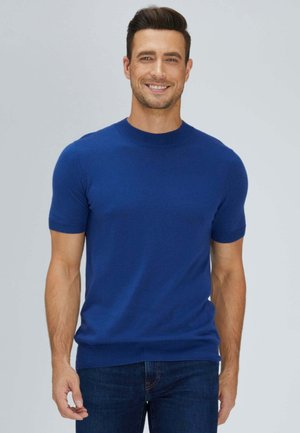 Lächelnder Mann mit kurzem dunklem Haar, der ein einfarbiges blaues kurzärmeliges Hemd und dunkelblaue Jeans vor einem hellgrauen Hintergrund trägt.