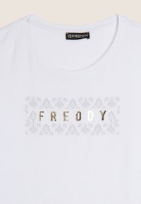 Camiseta blanca con cuello redondo que presenta un gráfico metálico plateado con la palabra "FREDDY" y un patrón decorativo en gris claro debajo del texto.