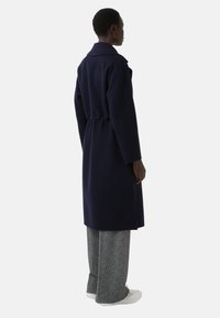 Marineblauwe wollen blend jas met een kraag, een getailleerde taille, lange mouwen en een knielange snit. Gecombineerd met grijs patroon brede broek.