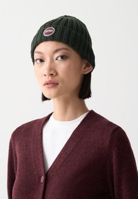 Căciulă tricotată verde închis cu textură ribbată și un patch circular maroniu. Asortată cu un cardigan burgundy peste o cămașă albă.
