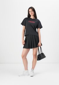 Black cotton t-shirt with red "Coca-Cola" logo, paired with a pleated black skirt, white high-top sneakers, and a small black handbag.