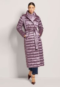 Lange pufferjas in lichtpaars, met een ceintuur in de taille, brede kraag en horizontaal gekwilt patroon. Draagt over een patroon top en jeans.