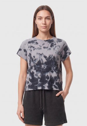 Mujer joven con cabello castaño liso que lleva una camiseta de tie-dye gris y negra y pantalones cortos oscuros de canalé con cordón.