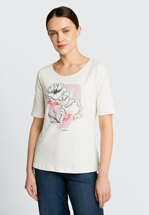 Kobieta ubrana w białą koszulę z krótkim rękawem z kwiatowym rysunkiem liniowym i pastelowymi blokami kolorów, zestawioną z ciemnoniebieskimi dżinsami.