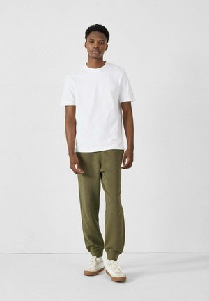T-shirt bianco in cotone abbinato a pantaloni della tuta verde oliva e sneakers bianche con suole beige. Il modello è in piedi contro uno sfondo neutro.