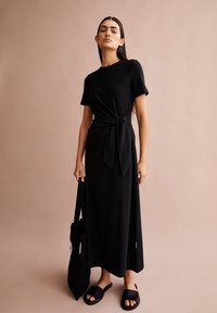 Schwarzes Kleid mit kurzen Ärmeln, knotet an der Taille, fließender Stoff, kombiniert mit einer schwarzen Tragetasche und dunklen Riemchensandalen vor einem beigen Hintergrund.