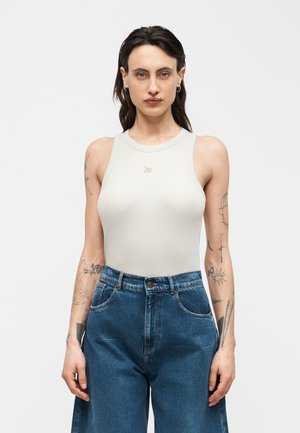 Donna con capelli scuri e tatuaggi che indossa un top bianco senza maniche a coste e jeans blu a vita alta su uno sfondo bianco semplice.