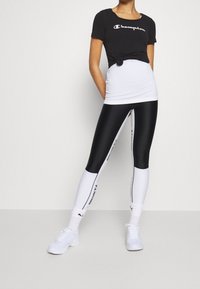 Černé těsné tričko s bílým logem, spárované s černobílými legínami se bočním textem. Model má na sobě bílé sportovní boty a ponožky.