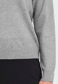 Grau gestrickter Pullover mit gerippten Bündchen und Saum, der eine glatte Textur und lange Ärmel aufweist, kombiniert mit schwarzen Hosen darunter.