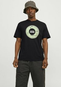 Zwarte katoenen t-shirt met een cirkelvormige groene graphic met de tekst "JACK & JONES" en "Expedition 99" op de borst, ronde halslijn, korte mouwen.