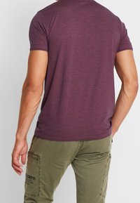 Camiseta corta de manga en color púrpura con cuello redondo, hecha de una tela suave, que presenta una pequeña etiqueta en el dobladillo, combinada con pantalones cargo en color verde oliva.