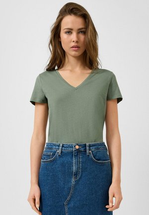 Young woman with loose light brown hair wearing a green V-neck T-shirt and high-waisted blue denim skirt standing against a light background.