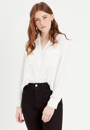 Camicia bianca a manica lunga con colletto, dotata di due tasche sul petto e una vestibilità ampia, abbinata a jeans neri a vita alta.