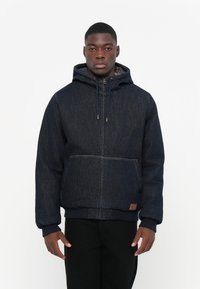 Temno jeans jakna s kapuco, z zadrgo in sprednjim žepom. Ima rebraste manšete in rob, z teksturirano tkanino in rjavim oznakim.
