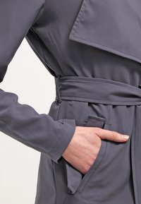 Szary płaszcz trencz z paskiem w talii, długimi rękawami i dużym kołnierzem. Po prawej stronie widoczna kieszeń. Gładka faktura.