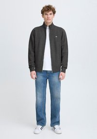 Kullsvart zip-up jakke med høy krage, parret med en hvit t-skjorte og lyseblå jeans. Modell som har på seg hvite joggesko.