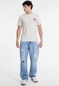Lichtbeige T-shirt met een ronde hals en een klein rood grafisch logo op de linkerborst, gecombineerd met lichtblauwe jeans met slijtage en witte sneakers.