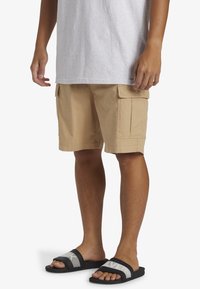 Shorts cargo beiges avec deux poches latérales et un tissu texturé. Portés avec des sandales à glissière noires présentant un logo gris.