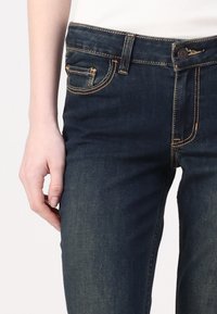 Σκούρες μπλε τζιν παντελόνες με αντίθετη πορτοκαλί ραφή, μπροστινή τσέπη και κούμπωμα. Χαρακτηρίζονται από λεία υφή και κομψή εφαρμογή.