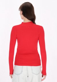 Maglione rosso a costine con maniche lunghe e collo alto, design aderente e texture liscia, abbinato a jeans chiari a vita alta.