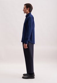 Camicia a maniche lunghe blu navy con bottoni, caratterizzata da un taglio dritto e fessure laterali, abbinata a pantaloni scuri e scarpe nere.