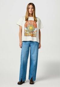 Camiseta gráfica crema que presenta un diseño de sol en naranja y azul. Combinada con jeans de pierna ancha azul claro con aberturas laterales y botas negras.