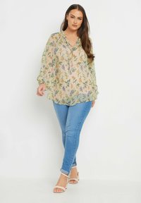 Blusa floreale trasparente con colori pastello, colletto abbottonato, polsini elasticizzati; abbinata a jeans skinny azzurri chiari e sandali beige.