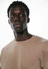 Hellbeige Pullover mit Rundhalsausschnitt und strukturiertem Strickmuster. Das Modell zeigt einen neutralen Gesichtsausdruck vor einem weißen Hintergrund.
