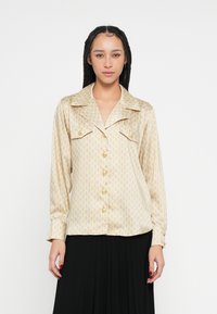 Blúzka s dlhým rukávom z béžového saténu so zlatým geometrickým vzorom, má vyrezávaný golier, predné vrecká a zlaté gombíky.