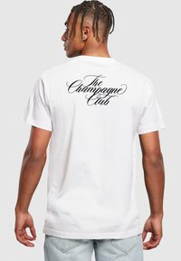 Camiseta de algodón blanca con un gran texto en cursiva negro "The Champagne Club" en la parte posterior. Mangas cortas y un corte relajado.