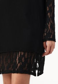 Zwarte jurk met een effen top en textuurkant mouwen. De kantzoom heeft een gedetailleerd patroon en eindigt net boven de pols.