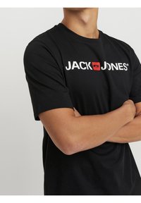 Camiseta de algodón negra con cuello redondo y mangas cortas. En la parte frontal muestra un texto de logo blanco "JACK & JONES" con un acento rojo.