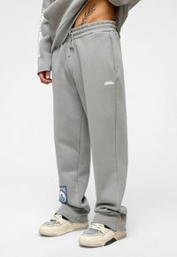 Grijze trainingsbroek met een elastische tailleband, zijzakken en een blauw logo-opdruk nabij de enkel. Gecombineerd met beige sneakers.